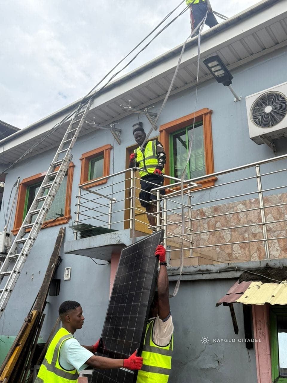 A Kayglo installer in a hi-vis vest passing a solar panel up from a balcony to the crew on the roof.